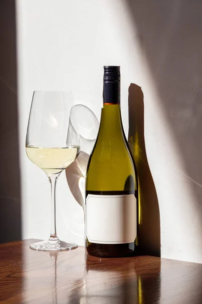 Bottle of white wine on the table, in the sunlight. Nearby is a glass of white wine. The bottle and glass stand on a varnished table and are reflected. A dark brown table stands against a white wall with Ukrainian ornaments.