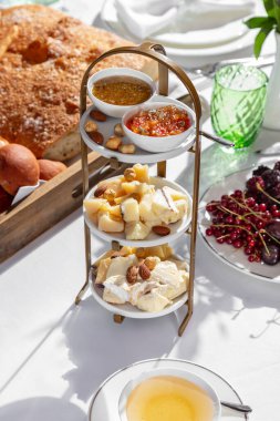 Beyaz tabaklarda, farklı seviyelerde, çeşitli peynir, fındık, bal ve tatlı sos çeşitleri vardır. Gövdeler metal bir büfede duruyor. Yakınlarda farklı ekmekleri olan bir tepsi, böğürtlenli bir tabak ve bir parça peynir görebilirsiniz..