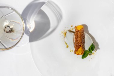 İçinde portakallı şerbet bulunan karamelli ve şam fıstıklı limonlu dondurma. Üstünde bir nane yaprağı var. Dondurma hafif, yuvarlak, geniş kenarlı seramik bir tabakta yatıyor. Tabak lambalı bir masanın üzerinde.