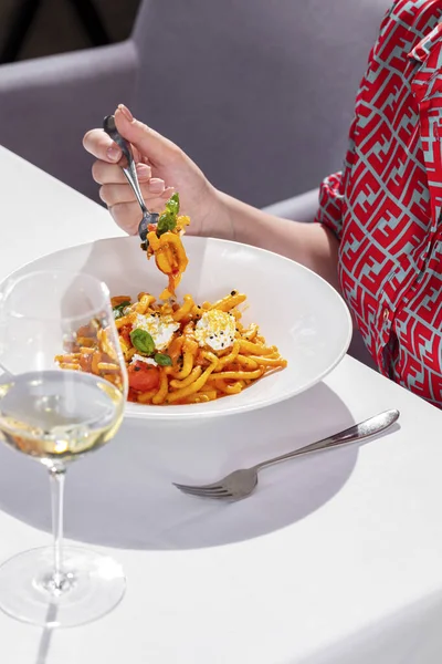 Domates soslu İtalyan makarna, fesleğen ve mozzarella. Makarna ışıkta seramik tabakta yatıyor. Çatalla makarna yiyen kız. Yakınlarda bir bardak beyaz şarap ve çatal bıçak takımı var. Masada beyaz bir masa örtüsüyle yemek ve şarap..