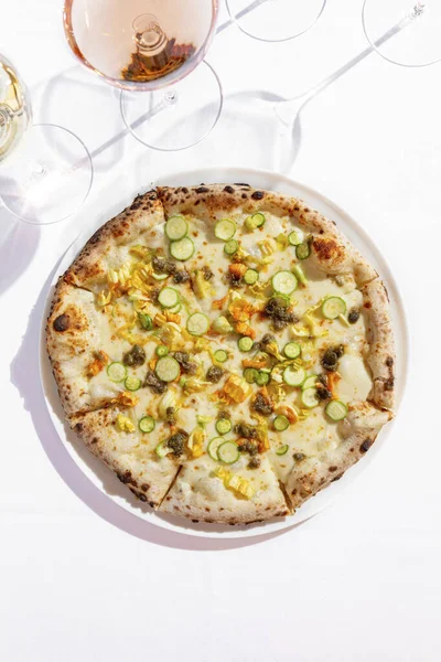 Italian vegetable pizza with zucchini, zucchini flowers, capers and cream sauce. Nearby are two glasses of rose and white wine. Hands take a piece on each side. Pizza and glasses are on a white tablecloth.
