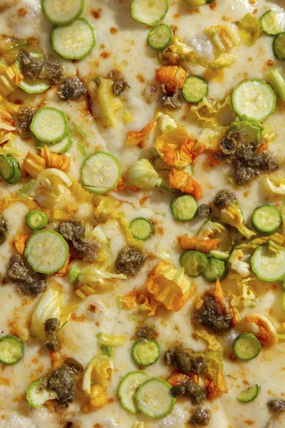 Italian vegetable pizza with zucchini, zucchini flowers, capers and cream sauce. Nearby are two glasses of rose and white wine. Hands take a piece on each side. Pizza and glasses are on a white tablecloth.