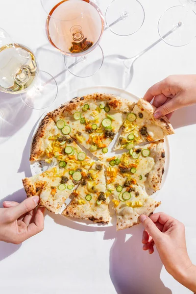 Italian vegetable pizza with zucchini, zucchini flowers, capers and cream sauce. Nearby are two glasses of rose and white wine. Hands take a piece on each side. Pizza and glasses are on a white tablecloth.