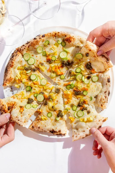 Italian vegetable pizza with zucchini, zucchini flowers, capers and cream sauce. Nearby are two glasses of rose and white wine. Hands take a piece on each side. Pizza and glasses are on a white tablecloth.