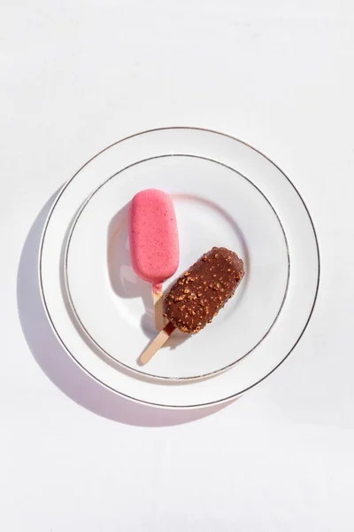 Chocolate glazed ice cream with nuts on a wooden stick and strawberry glazed ice cream on a wooden stick. Ice cream lies on a round, ceramic plate with a tarp. Dishes are on a white tablecloth.