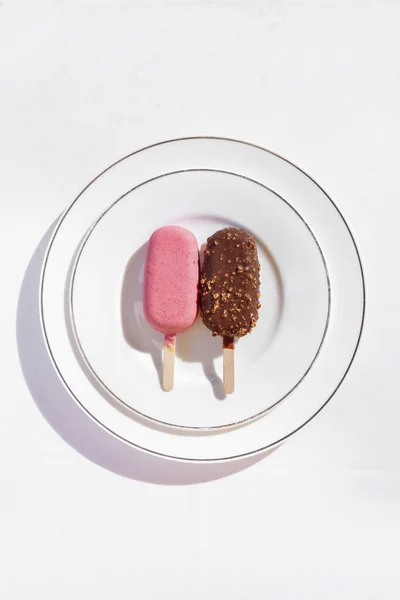 Chocolate glazed ice cream with nuts on a wooden stick and strawberry glazed ice cream on a wooden stick. Ice cream lies on a round, ceramic plate with a tarp. Dishes are on a white tablecloth.