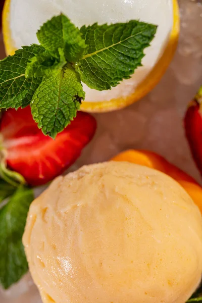 Yuvarlak bir tabakta, 6 farklı çeşit dondurma ve çikolatalı dondurma var. Dondurma küçük bir buz kırıntısının üzerinde, yakınlarda böğürtlenler var. Tabak beyaz bir masa örtüsünün üzerinde..