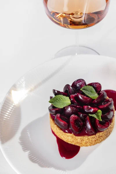 Cherry tart with fresh cherries, buttercream and shortcrust pastry. Tart lies on a white plate, with cherry sauce. Nearby is a glass of rose wine. A plate and a glass stand on a white tablecloth.