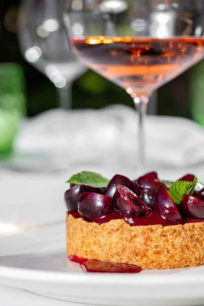 Cherry tart with fresh cherries, buttercream and shortcrust pastry. Tart lies on a white plate, with cherry sauce. Nearby is a glass of rose wine. A plate and a glass stand on a white tablecloth.