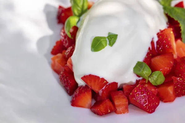 Vanilla ice cream with strawberries and basil. A portion of vanilla ice cream lies on top of finely chopped strawberries. Nearby lies a sprig of mint and basil leaves. Ice cream lies in a white, round, ceramic plate with wide margins. The plate stand