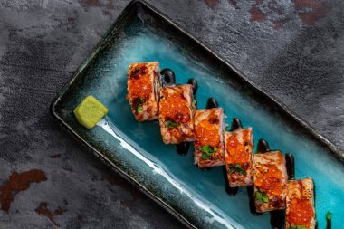 Sushi wrapped in salmon fillet, with red caviar on top and finely chopped green onions, stuffed with avocado and eel in unagi sauce. Sushi stands on a blue, rectangular, ceramic plate with dark sides. Unagi sauce is poured on a plate, there is a squa
