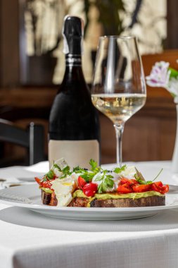 Italian dish. Buckwheat bread bruschetta with mashed avocado, cherry tomatoes, artichokes, parsley leaves and two poached eggs topped with ground truffle. Bruschetta stands on a round, white ceramic plate. Nearby is a champagne glass with a long stem