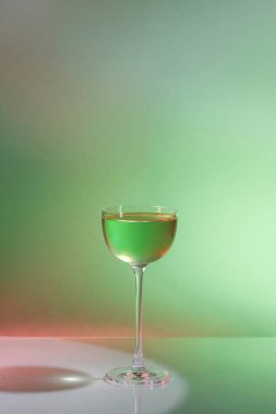 Gin cocktail Matrix. A gin-based cocktail, green in color, poured into a vermouth glass with a tall glass stem. Next to the glass is a beaker with a transparent liquid and green paint dissolves in it. A glass and a beaker stand on a glass background.