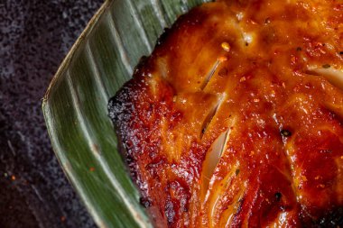 Grilled sea bass fillet in unagi sauce. The fillet lies on a palm leaf. Leaf and fish lie on a ceramic square plate. The plate stands on a gray stone background with bronze inlays.