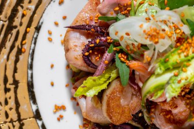 Deniz mahsullü sıcak salata. Ahtapot salatası, kalamar, karides, istiridye, paprika içinde susam tohumu, Japon mayonezinde marul ve tartar sosu. Salata, geniş kenarlı altın bir tabakta yatıyor. Plaka siyah bir kağıdın üzerinde duruyor ve karoların üzerinde g harfi var.