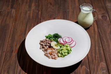 Domuz parçaları, yumurtalı tavuk, salatalık, soğan, turp ve maydanoz ahşap arka planda bir tabakta Okroshka ile sürahide.