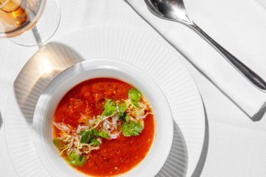 Spanish gazpacho soup with crab meat and greens in a deep, light plate. The plate is on a table with a light tablecloth, next to it is a glass of rose wine and a bottle of olive oil, and cutlery.