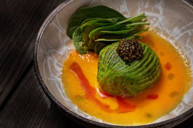 Scallop sashimi under a pillow of avocado slices, topped with a handful of black caviar. Sashimi in yellow tomato sauce with paprika, lettuce sprinkled with turmeric. The dish is in a round, ceramic, brown bowl with a pattern. The plate stands on a b