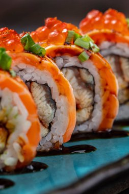 Sushi wrapped in salmon fillet, with red caviar on top and finely chopped green onions, stuffed with avocado and eel in unagi sauce. Sushi stands on a blue, rectangular, ceramic plate with dark sides. Unagi sauce is poured on a plate, there is a squa