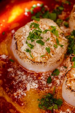 Sea scallop fried in white wine with tomato sauce and wig, finely chopped cilantro. The food lies in a ceramic, blue plate with high sides. The plate stands on a dark, wooden background.