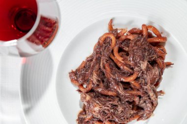 Italian dish. Pasta Bucatini with finely chopped duck fillet in plum sauce. The pasta lies in a deep, round, wide-brimmed ceramic plate. The plate is on the table on a white tablecloth. Next to the plate is a long-stemmed glass with red wine, a bowl 