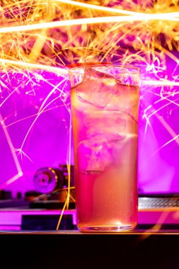 Lemonade and ice are poured into a tall and transparent glass. Around the glass is a drawing on a long exposure, with Bengal fire. A part of a vinyl player is visible next to the glass.