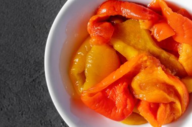 İtalyan otları ve sarımsakla marine edilmiş kırmızı ve sarı biber. Paprika beyaz, yuvarlak, seramik bir kasede yatıyor. Kase gri, taş bir arka planda duruyor..