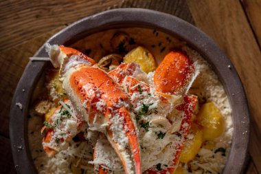 Potatoes in cream cheese sauce and white wine with crab meat. Sprinkle generously with Parmesan cheese on top. The dish lies in a round, ceramic plate. The plate is on a wooden table with a pattern. Nearby is a glass of white wine, a salt and pepper 