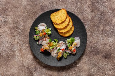 Uskumru ve ringa filetosu, ince doğranmış sebzeler, mikroyeşil lahanalar ve mantar sosuyla dilimlenmiş. Balık siyah, yuvarlak, tahta tabakta yatıyor. Yakınlarda kızarmış mısır ekmeği var. Tabak koyu renk, kumaş arka plan, kahve. 