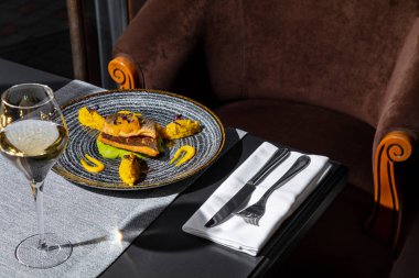 Fried fish with mashed potatoes, sprouts, mango, and sauce in a plate on the table with a napkin, cutlery and a glass of wine