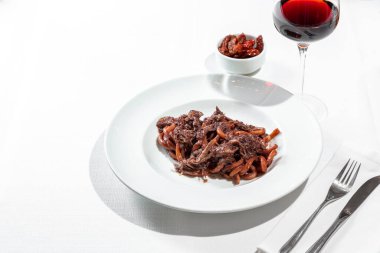 Italian dish. Pasta Bucatini with finely chopped duck fillet in plum sauce. The pasta lies in a deep, round, wide-brimmed ceramic plate. The plate is on the table on a white tablecloth. Next to the plate is a long-stemmed glass with red wine, a bowl 