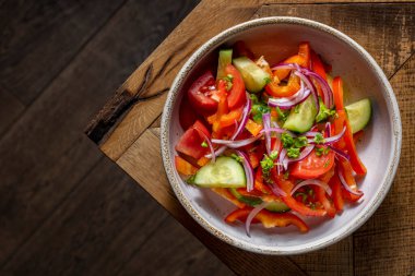 Spring salad with fresh vegetables. Salad with fresh tomatoes, paprika, cucumber, red onion, chopped cilantro and olive oil. The salad lies in a round, deep, ceramic bowl. The dishes are on a wooden table with a pattern.