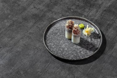 Üzerinde wagyu bifteği ve mikroyeşil soğan filizi olan ince pirinç hamuruna sarılmış pirinç gunkan. Wagyu 'lu iki silahşör gri, yuvarlak, düşük kenarlı seramik bir tabakta yatıyor. Tabak gri bir arka planda duruyor..