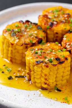 Çeyrekliklere bölünmüş ve ızgara yapılmış bir mısır, bir tabakta yatıyor ve üzerine sarı mango sosu serpilmiş, ince doğranmış yeşil soğan ve zerdeçal serpiştirilmiş. Mısır yuvarlak, hafif seramik bir tabakta yatıyor. Tabak bej tuğlalı arka planda duruyor..