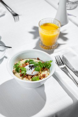 Breakfast. Morning dish of salted oatmeal with almonds, poached eggs and parsley leaves. Porridge with ingredients lies in a white, ceramic, deep bowl. The bowl is on a table with a white tablecloth. Next to the porridge is a glass of orange juice an