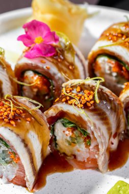 Pan-Asya mutfağı, unagi soslu yılan balığı, pirinç, salatalık ve Philadelphia peyniri. Susam tohumları ve mikroyeşil lahanalarla kaplı. Suşi yuvarlak, hafif, düşük kenarlı seramik bir tabakta yatar. Yakında zencefil turşusu ve wasabi küpü var. Tabak Stan.