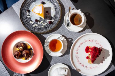 Cheesecake with jam, cream, blackberries and slices in the first plate, a piece of cake with cream, cherries and cherry jam in the second plate, profiteroles with chocolate crumbs, chocolate plates, blackberries, cream and bread on the table with cup