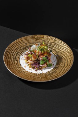Deniz mahsullü sıcak salata. Ahtapot salatası, kalamar, karides, istiridye, paprika içinde susam tohumu, Japon mayonezinde marul ve tartar sosu. Salata, geniş kenarlı altın bir tabakta yatıyor. Tabak siyah bir arka planda duruyor..