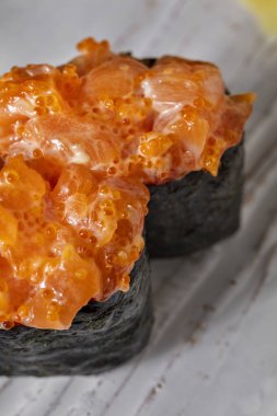 Gunkan with salmon, Japanese chili mayonnaise and tobiko caviar. Two gunkans stand on a rectangular, light, ceramic plate. The plate is on a wooden table with a pattern.