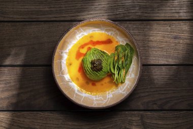 Scallop sashimi under a pillow of avocado slices, topped with a handful of black caviar. Sashimi in yellow tomato sauce with paprika, lettuce sprinkled with turmeric. The dish is in a round, ceramic, brown bowl with a pattern. The plate stands on a b