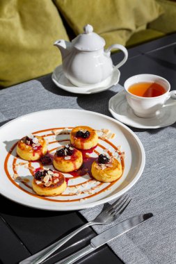 Cheesecakes with jam, blackberries and almond slices in a plate on the table with a cup of tea, teapot, cutlery, and a vase with a flower