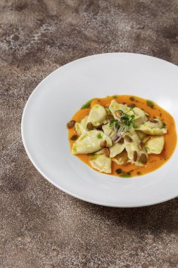 Porcini köftesi, kabak sosu, kabak çekirdeği, balzamik pesto sosu, mikroyeşil lahanalar. Yiyecek beyaz, yuvarlak, derin, seramik bir tabakta yatıyor. Tabak koyu renk, kumaş arka plan, kahve rengi..