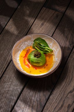 Scallop sashimi under a pillow of avocado slices, topped with a handful of black caviar. Sashimi in yellow tomato sauce with paprika, lettuce sprinkled with turmeric. The dish is in a round, ceramic, brown bowl with a pattern. The plate stands on a b