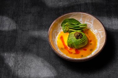 Scallop sashimi under a pillow of avocado slices, topped with a handful of black caviar. Sashimi in yellow tomato sauce with paprika, lettuce sprinkled with turmeric. The dish is in a round, ceramic, brown bowl with a pattern. The plate stands on a g