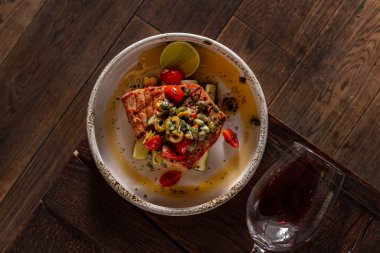 Tuna fillet steak, on fried zucchini wedges, with olive salsa on top. Olive oil with herbs is spilled nearby and a lime wedge lies. The steak lies in a white, round, ceramic plate with high sides. The plate stands on a wooden table with wood texture 