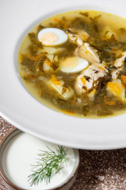 Beyaz, yuvarlak, derin, seramik tabakta yeşil borş çorbası, haşlanmış yumurta, haşlanmış domuz eti, havuç ve patates. Tabak koyu kahve arka planında duruyor. Yakınlarda küçük, beyaz, ekşi kremalı seramik bir kase var, üzerinde maydanoz var.