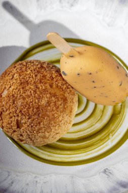 Italian dessert. In a deep, handmade ceramic plate, there is a cake with vanilla cream and coffee ice cream. Pistachio sauce is smeared on the bottom of the plate. The plate is on a table with a white tablecloth. Nearby is a glass with homemade tinct