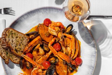 Langoustines, salmon, mussels, stewed tomatoes and shrimps in tomato sauce in a large ceramic plate. Nearby are two slices of buckwheat bread. A plate stands on a table with a light tablecloth, two glasses of rose wine stand nearby, and cutlery lies.
