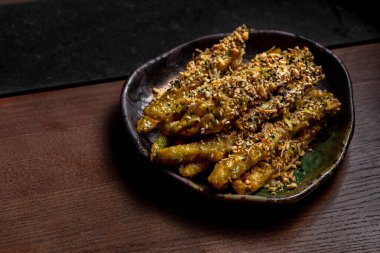 Deep-fried asparagus with sweet and sour sauce and sprinkled with sesame seeds with crushed cashews. The food is in a ceramic deep plate. The plate stands on a wooden background.