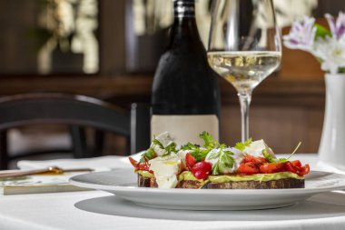 Italian dish. Buckwheat bread bruschetta with mashed avocado, cherry tomatoes, artichokes, parsley leaves and two poached eggs topped with ground truffle. Bruschetta stands on a round, white ceramic plate. Nearby is a champagne glass with a long stem